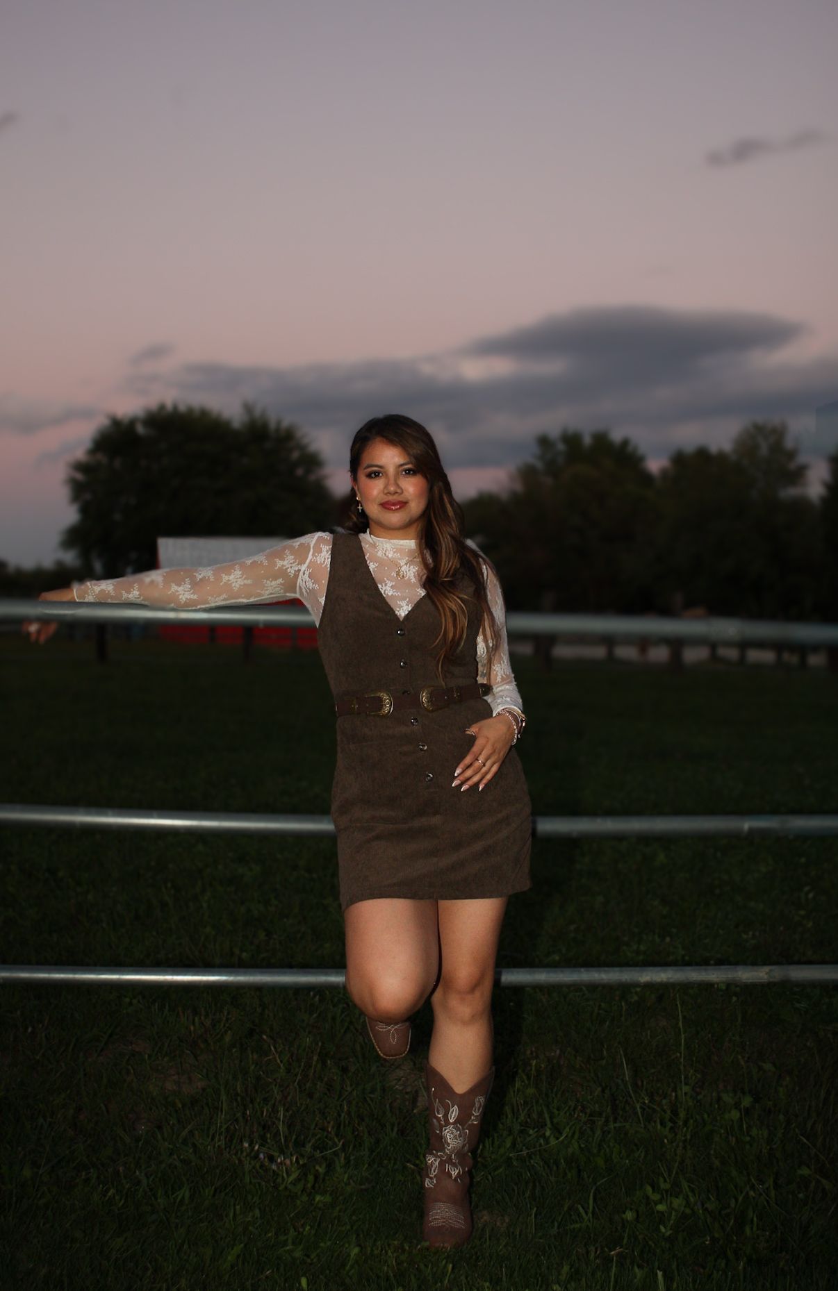 Person in brown dress and boots poses against sunset backdrop in countryside setting.