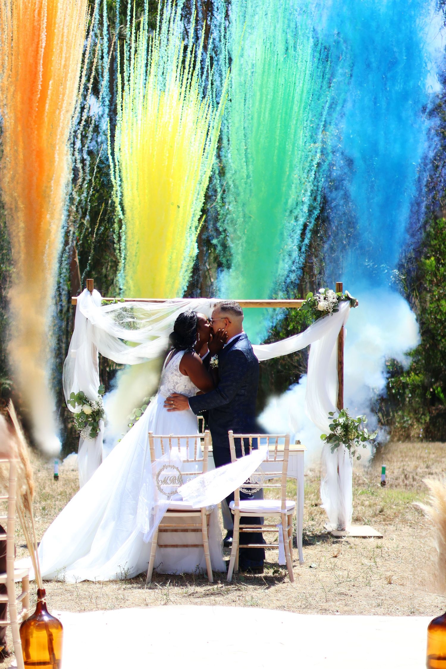 Wedding ceremony under colorful smoke effects with white draped arch and rustic decor.