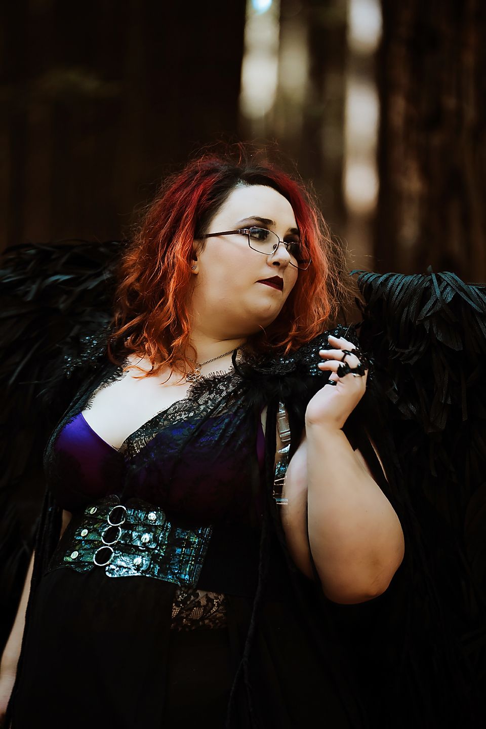 Gothic model in black corset and wings poses dramatically in moody forest setting with red hair and glasses.