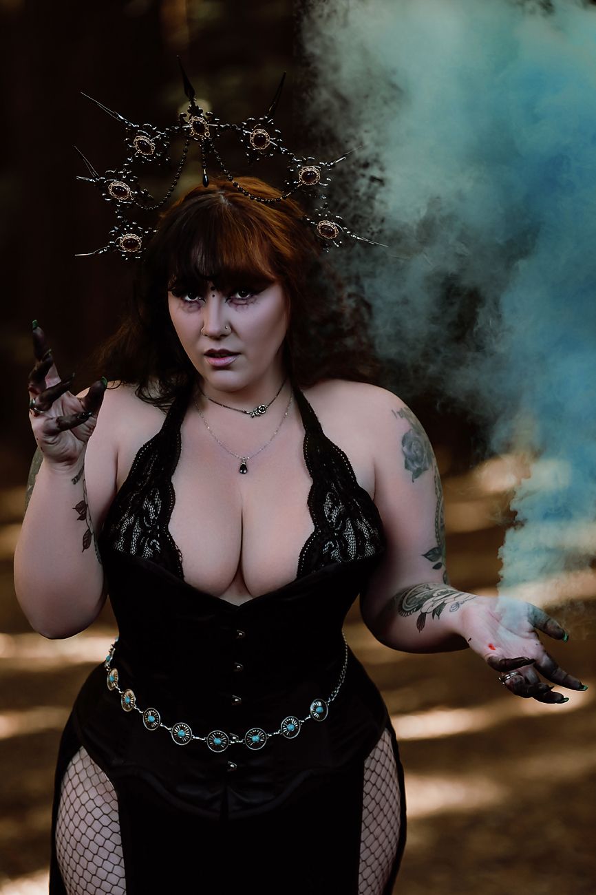 Dramatic forest photoshoot featuring gothic model in black lace and ornate crown with blue smoke effects.