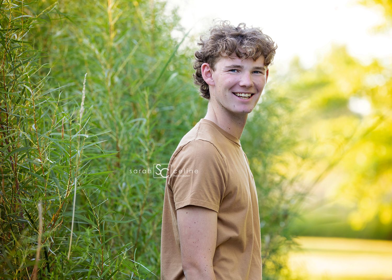 Person wearing tan shirt smiling warmly while standing in front of sun-dappled greenery in outdoor setting.