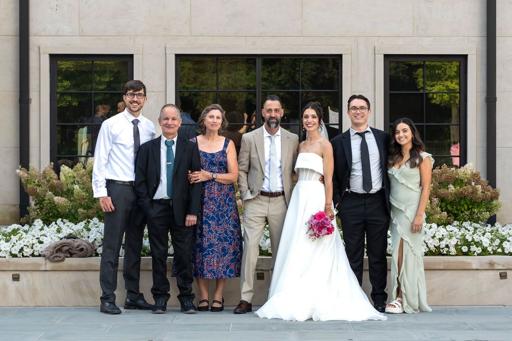 Wedding party members pose together in formal attire outside a building with white flowers in the background.