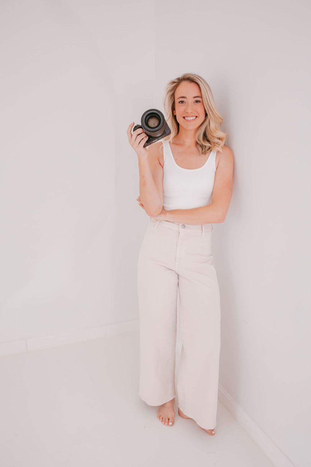 Photographer standing barefoot in white studio holding camera lens with genuine smile.