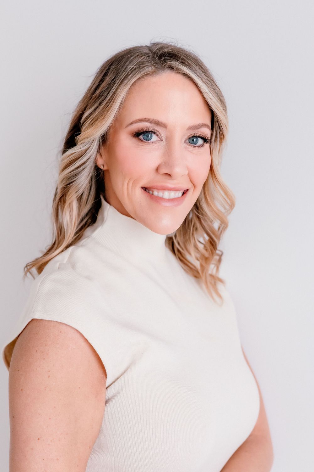 Professional portrait of person wearing cream turtleneck sleeveless top against light background.