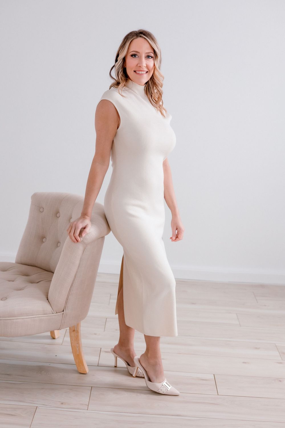 Full-length portrait in cream turtleneck dress standing next to beige chair.