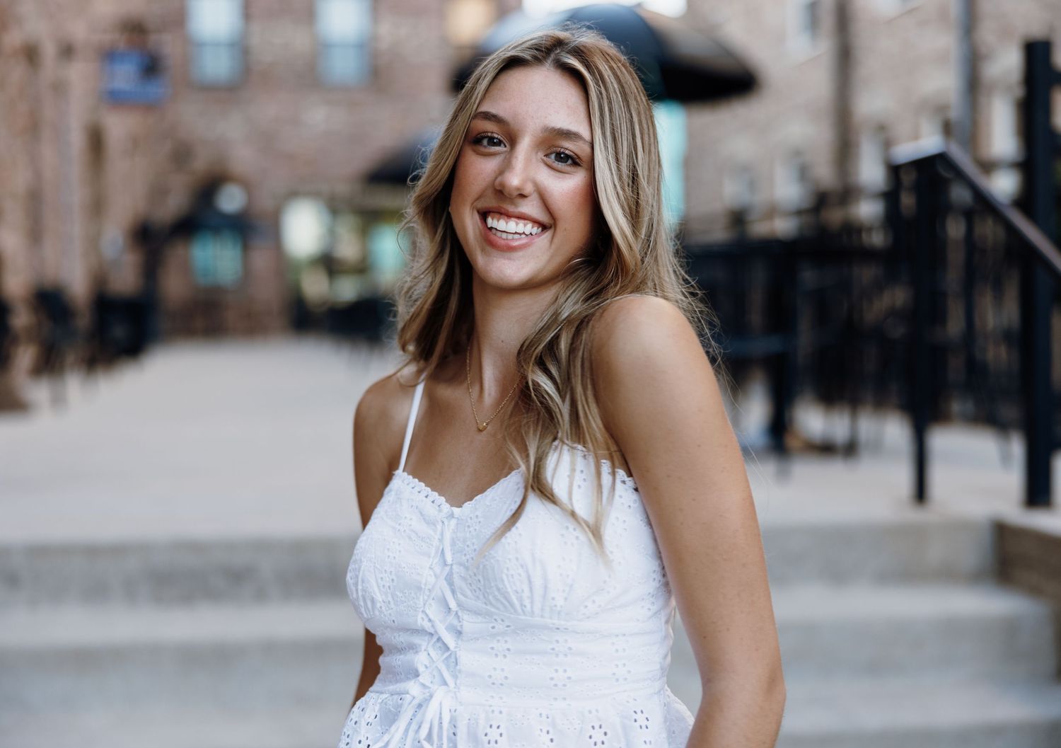Woman in white dress smiling against urban brick building backdrop.