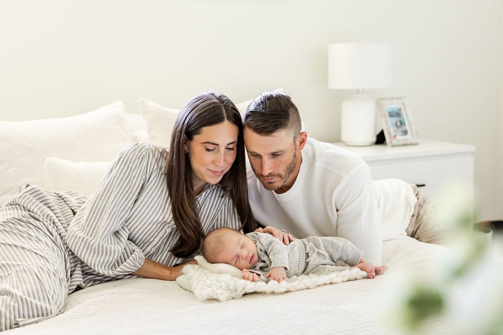 Parents in white outfits share tender moment with newborn baby against neutral backdrop in studio setting.