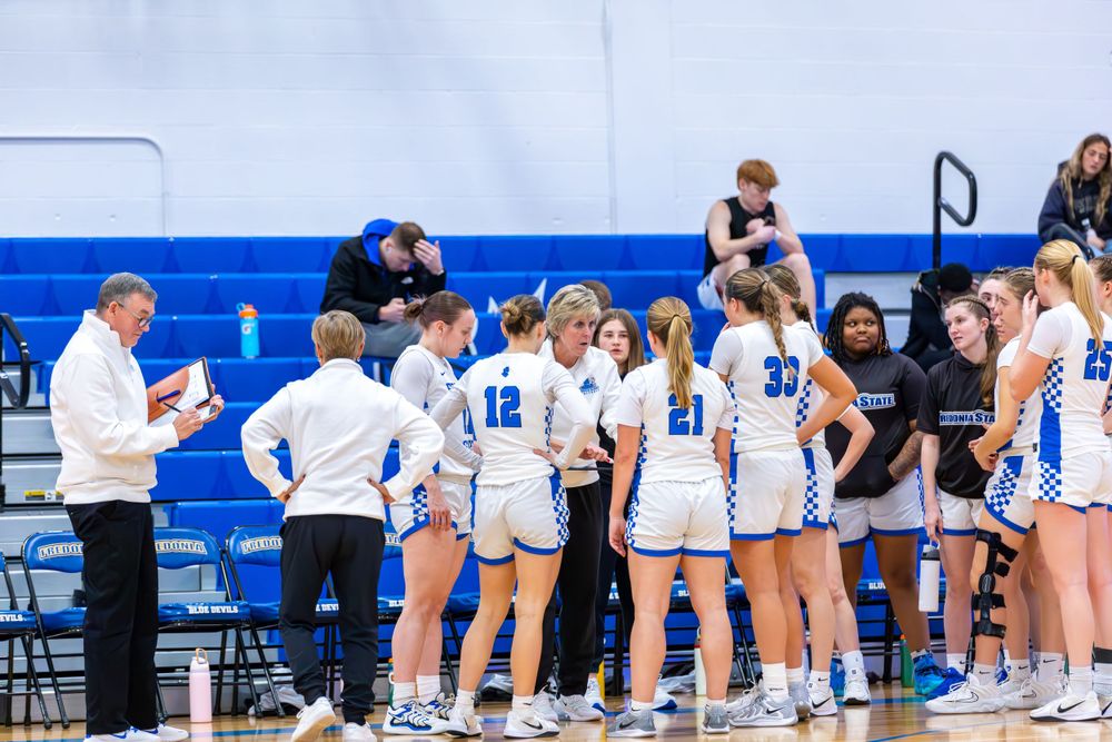 Basketball teams line up along the sidelines of a blue and white gymnasium during a game.