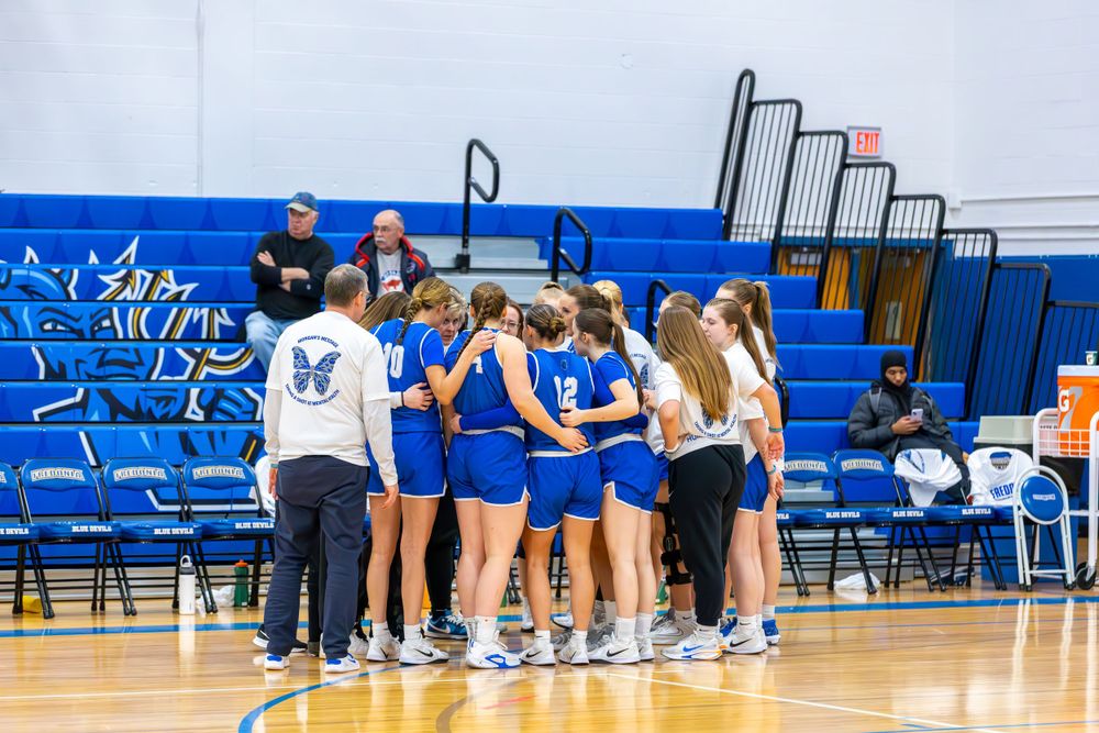 Team members line up on chairs along the sideline of a blue-themed basketball court.