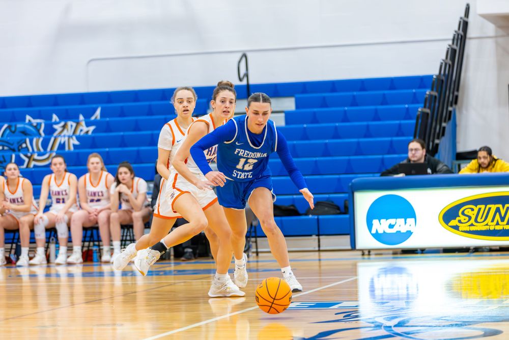 Players reach for a basketball during a competitive match in a gymnasium with NCAA banners.