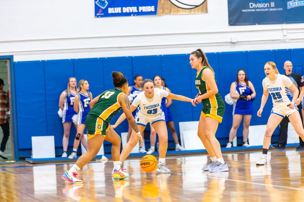 Players dive for a loose ball during an intense basketball game in a blue-walled gymnasium.