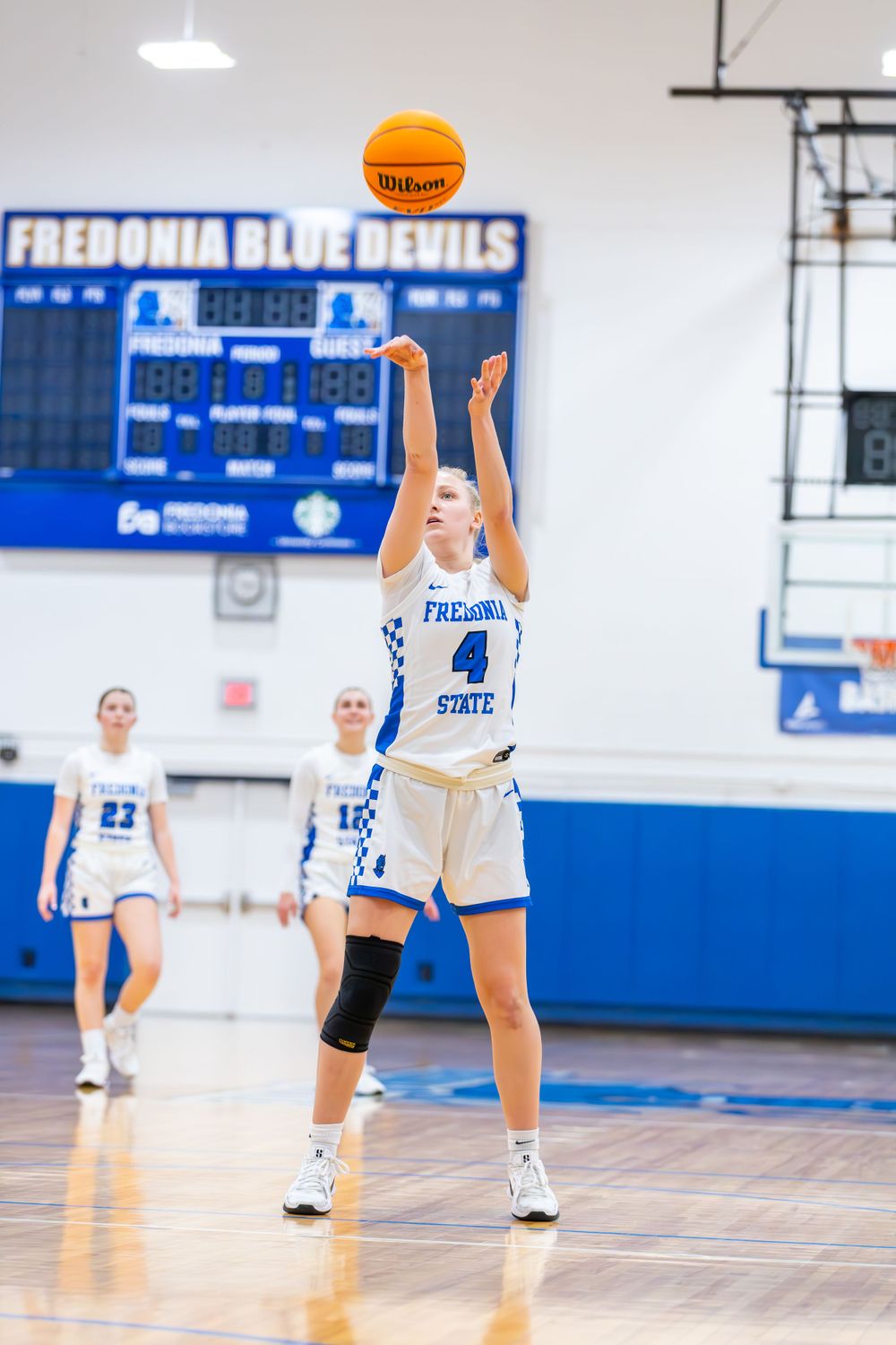 Players in white uniforms practice shooting drills on a basketball court with blue accents.