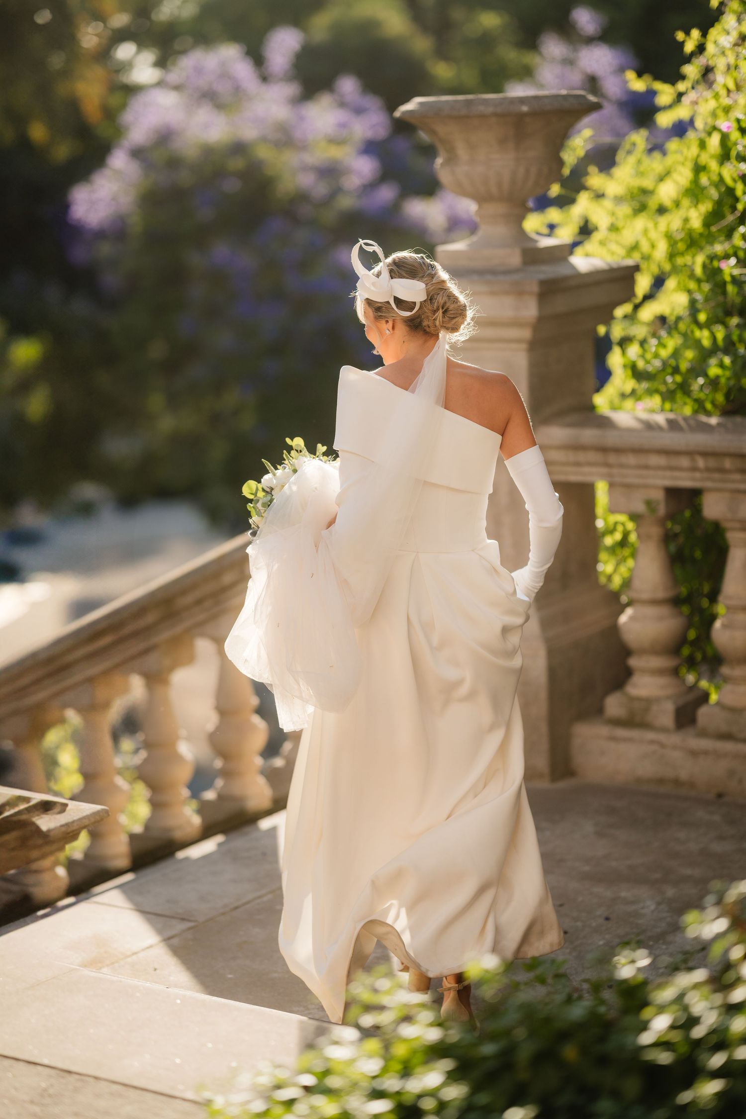 Elegant bride in white structured gown with gloves walks along stone balustrade in sunlit garden.