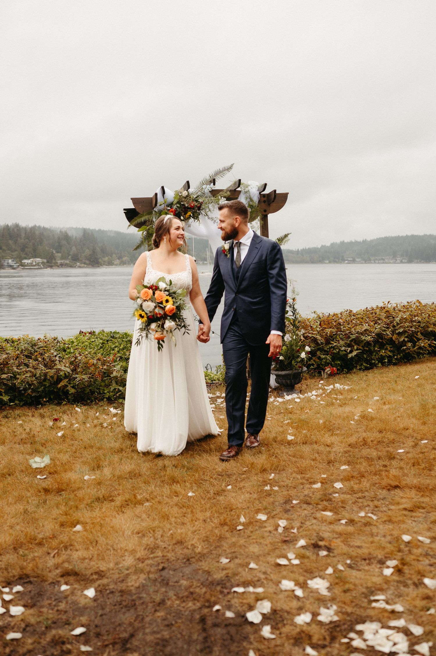 Wedding couple walking on coastal path with rustic wooden arch and water view.