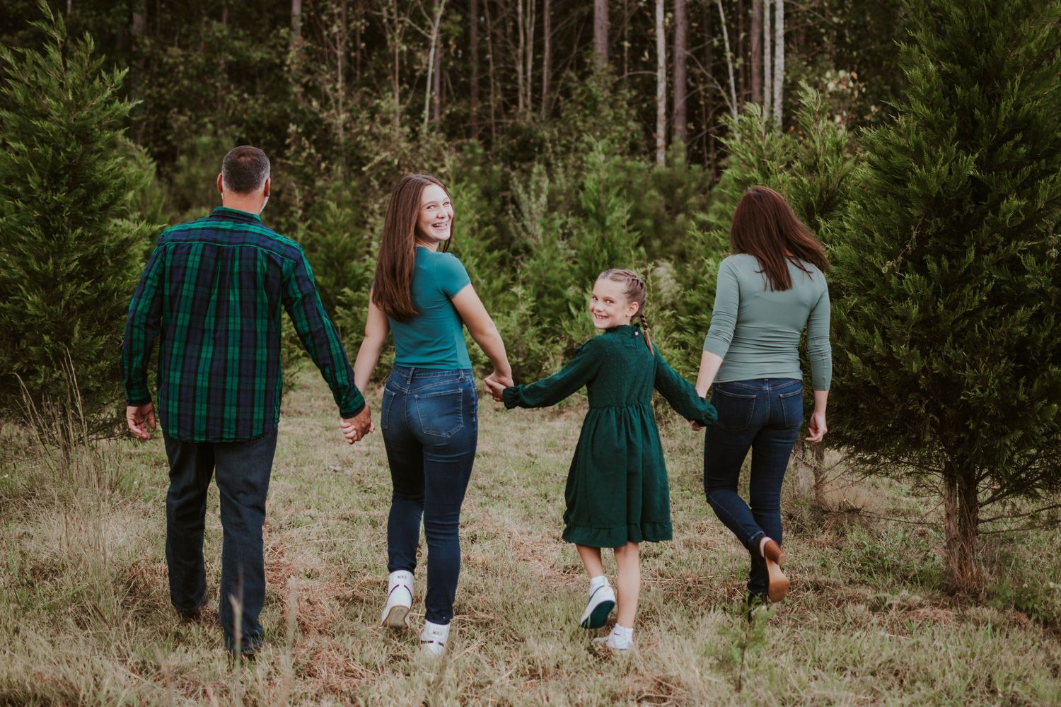 A family holds hands while walking together through a Christmas tree farm wearing coordinated green outfits.
