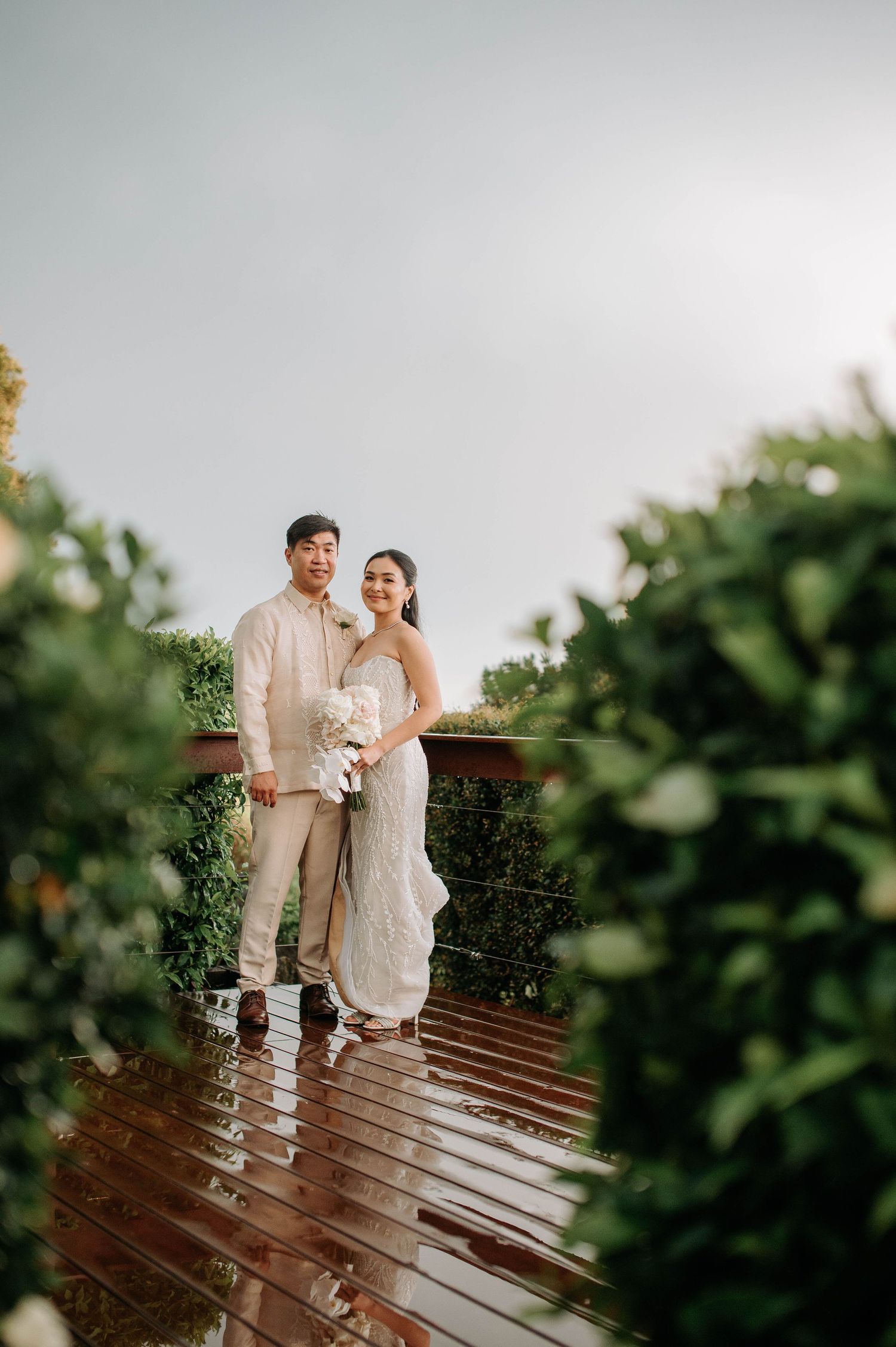 Wedding couple standing on wet wooden deck surrounded by lush greenery.