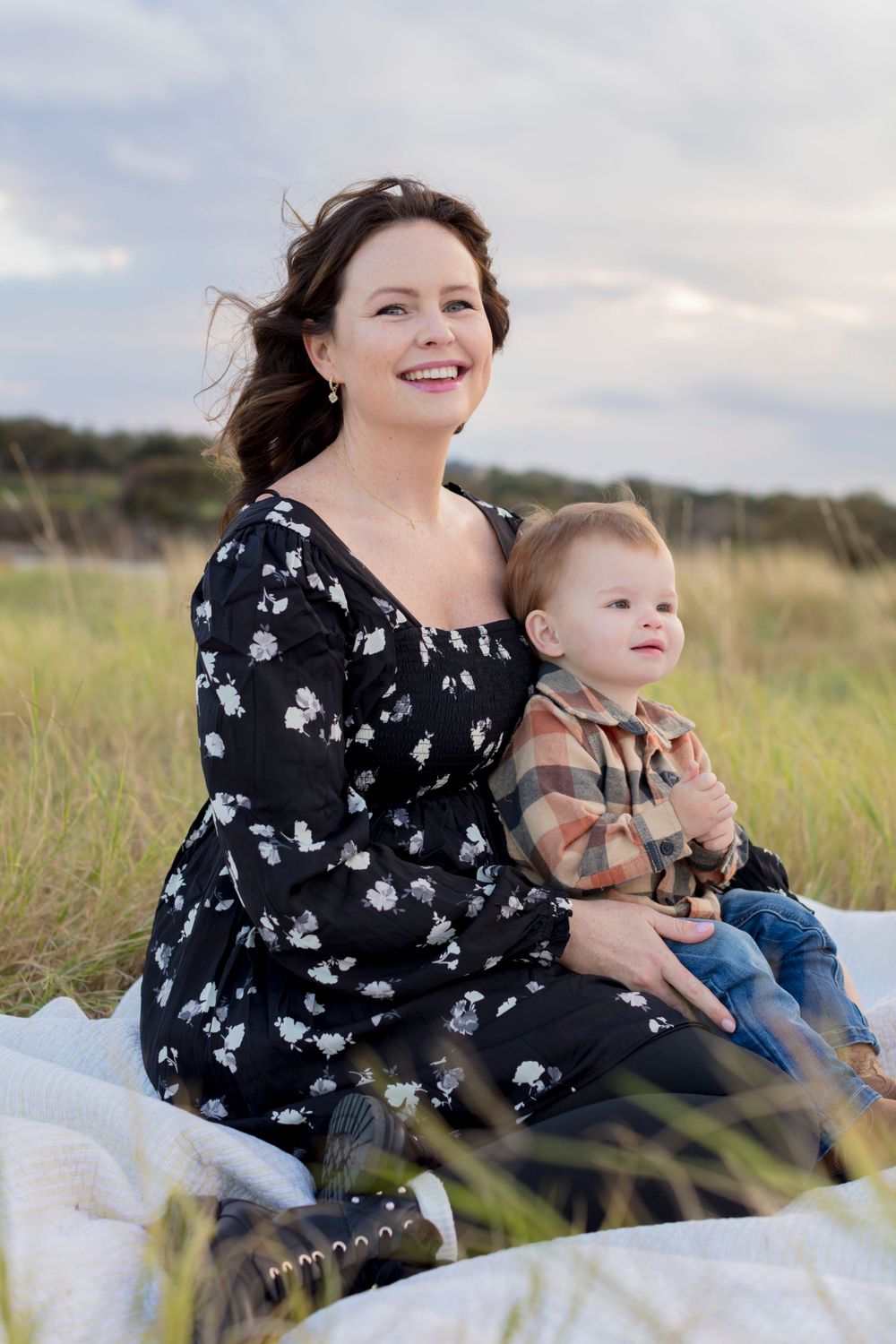 A parent and young toddler sit together on a blanket in a sunny field with tall grass swaying in the breeze. Cedar Park Mini Session