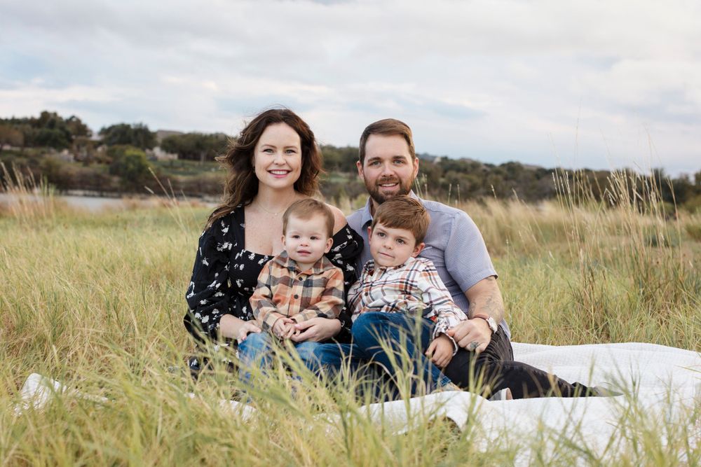 A family sits together on a blanket in a grassy meadow on an overcast day. Fall family Mini Session Cedar Park Texas