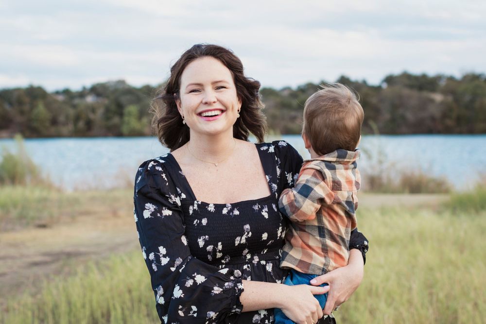 Person in a floral dress holding a child in plaid against a lakeside backdrop.