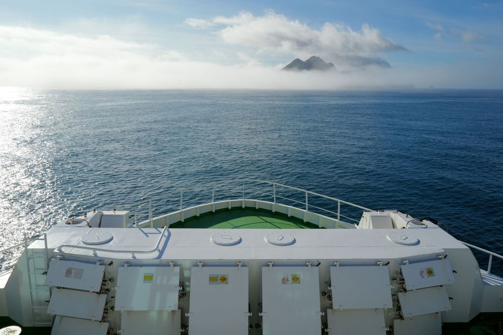 View from cruise ship deck showing railing and bow looking out towards misty mountains on horizon over blue ocean waters.