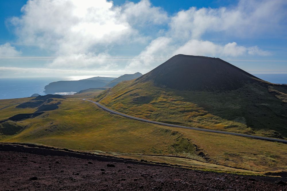 Winding coastal road curves around verdant hills and volcanic peaks on a remote island under dramatic clouds.