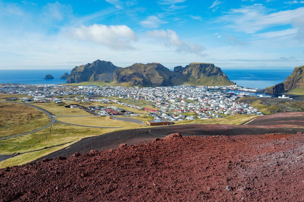 Coastal town Heimaey nestled between volcanic mountains and ocean on Vestmannaeyjar island in Iceland with red soil slopes.