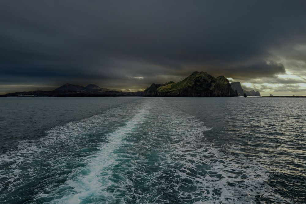 Dramatic stormy skies over rocky coastal cliffs with boat wake trailing through dark ocean waters.
