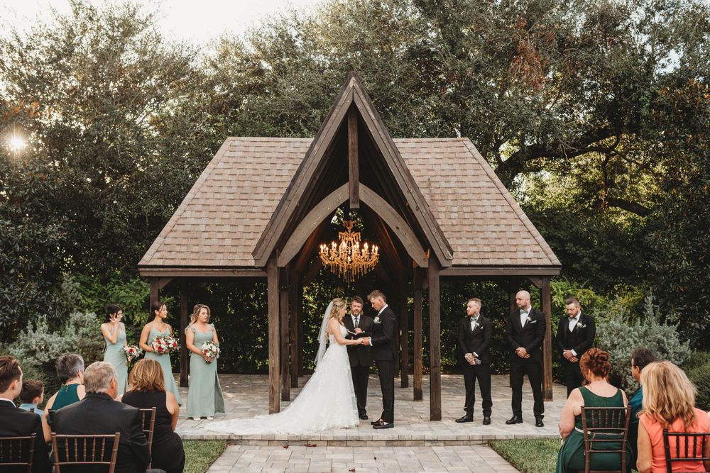 Wedding ceremony under wooden chapel with chandelier and guests seated outdoors.