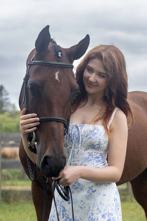 A person in a floral dress poses with a brown horse in a grassy field on an overcast day.