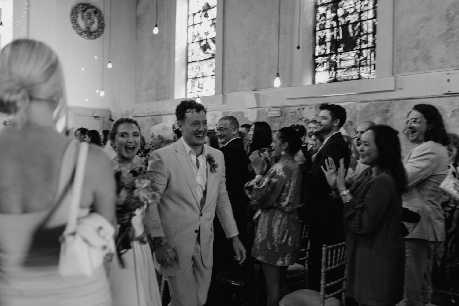 Wedding guests applaud during a joyous moment inside a church with stained glass windows in the background.