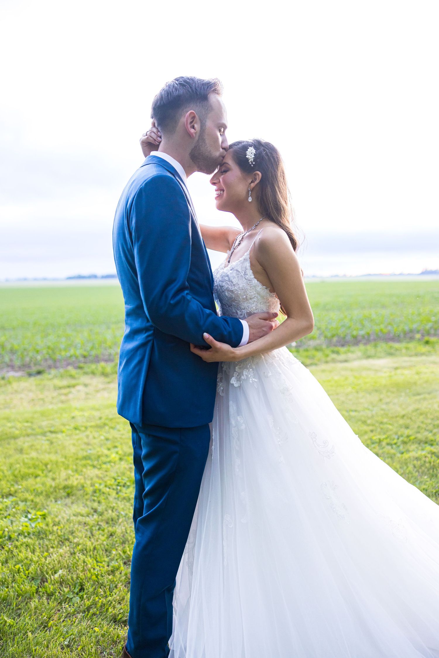 A couple in wedding attire embraces in an open field with dramatic sky backdrop.