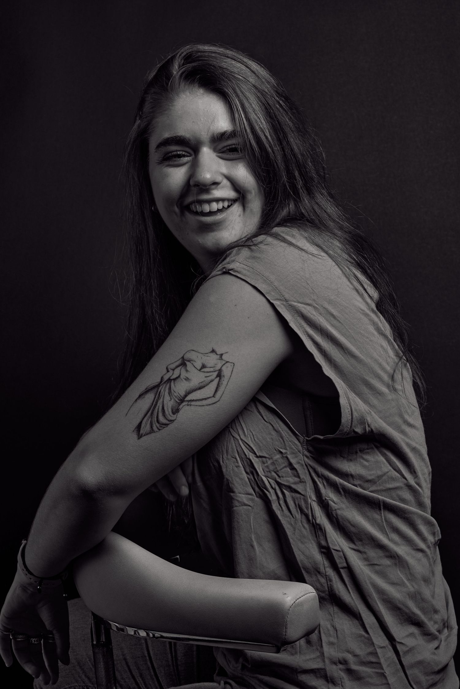 Zwart wit foto van een jonge vrouw met tattoos die lacht terwijl ze op een stoel zit.