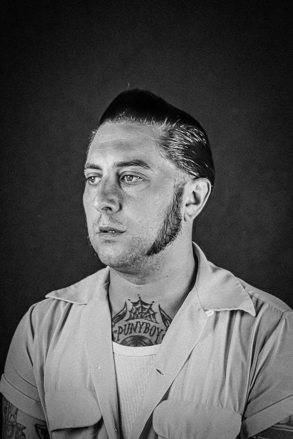 Black and white portrait of a person in a light jacket with neck tattoo and slicked back hair looking to the side.