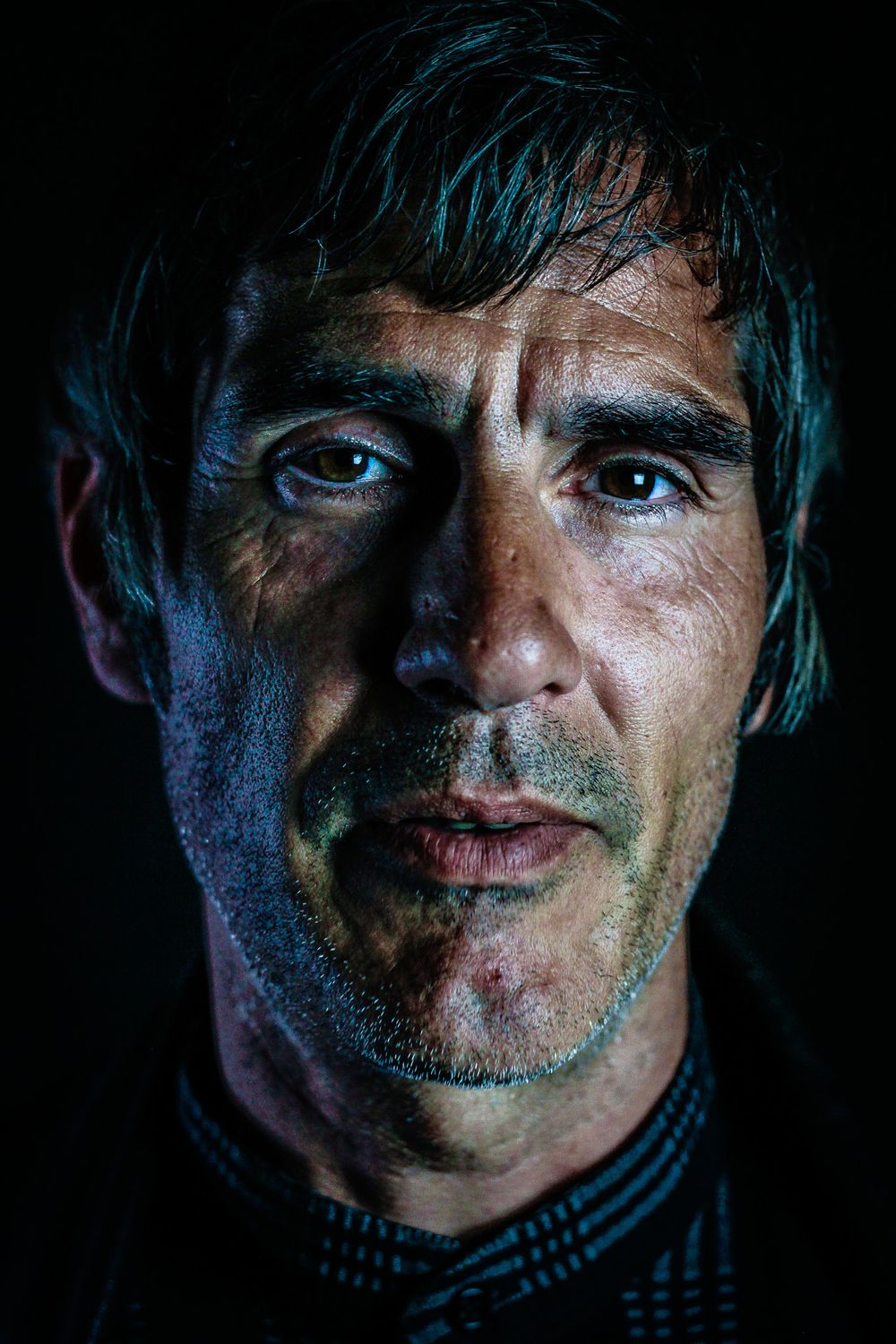 Dramatic close-up portrait with blue lighting shows intense eyes and textured skin against dark background.