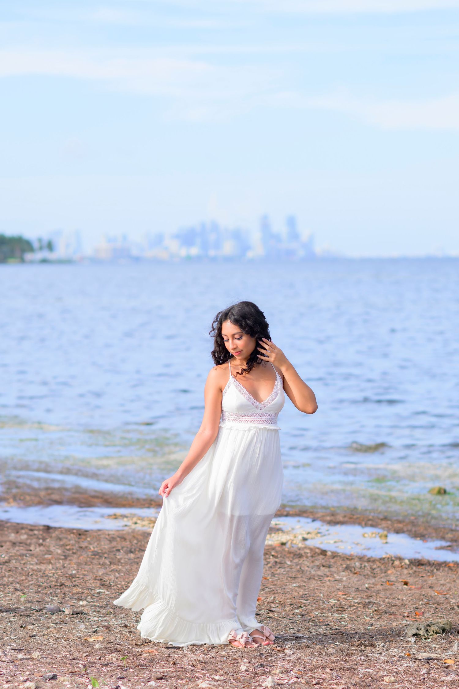 A teen girl in a flowing white maxi dress poses on a beach with a city skyline visible in the distance.
