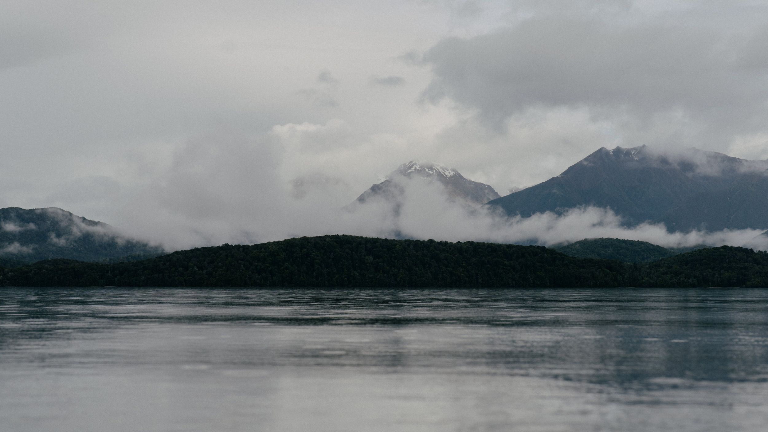 Misty mountains and forests reflect in still lake waters under cloudy skies.