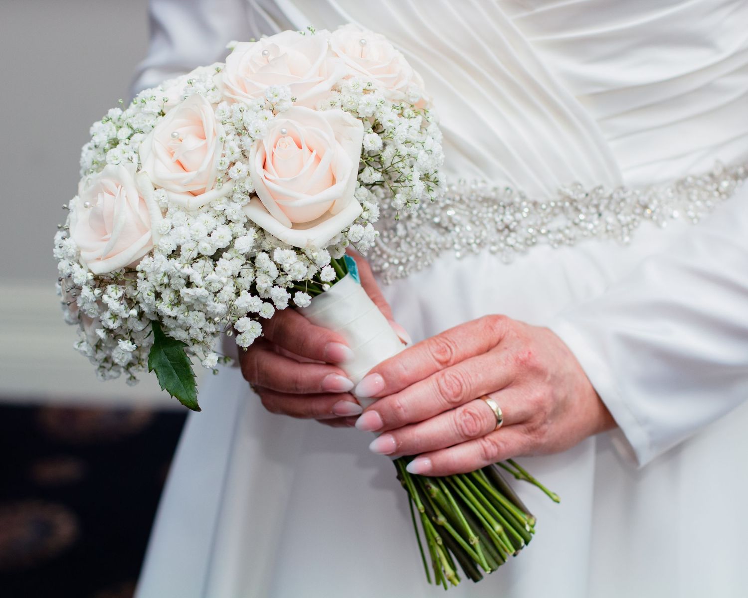 A wedding bouquet with soft pink roses and baby's breath held by hands in white sleeves.