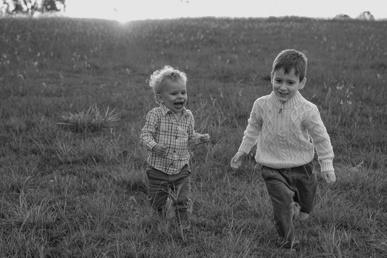 Black and white photo of two laughing young brothers running together across a grassy field in warm evening light.