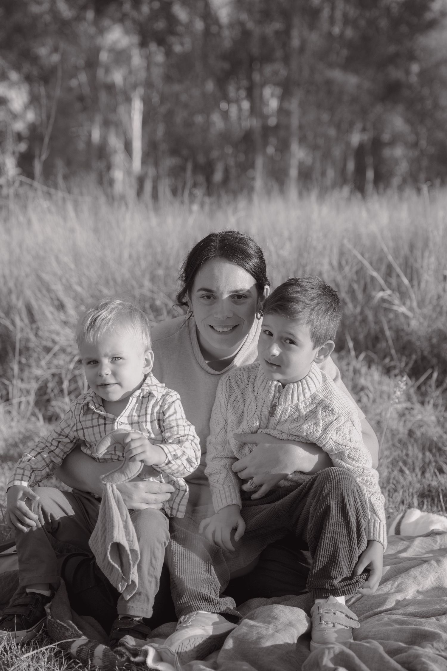 Black and white portrait of a smiling mother seated on a blanket in tall grass, holding her two young sons close.