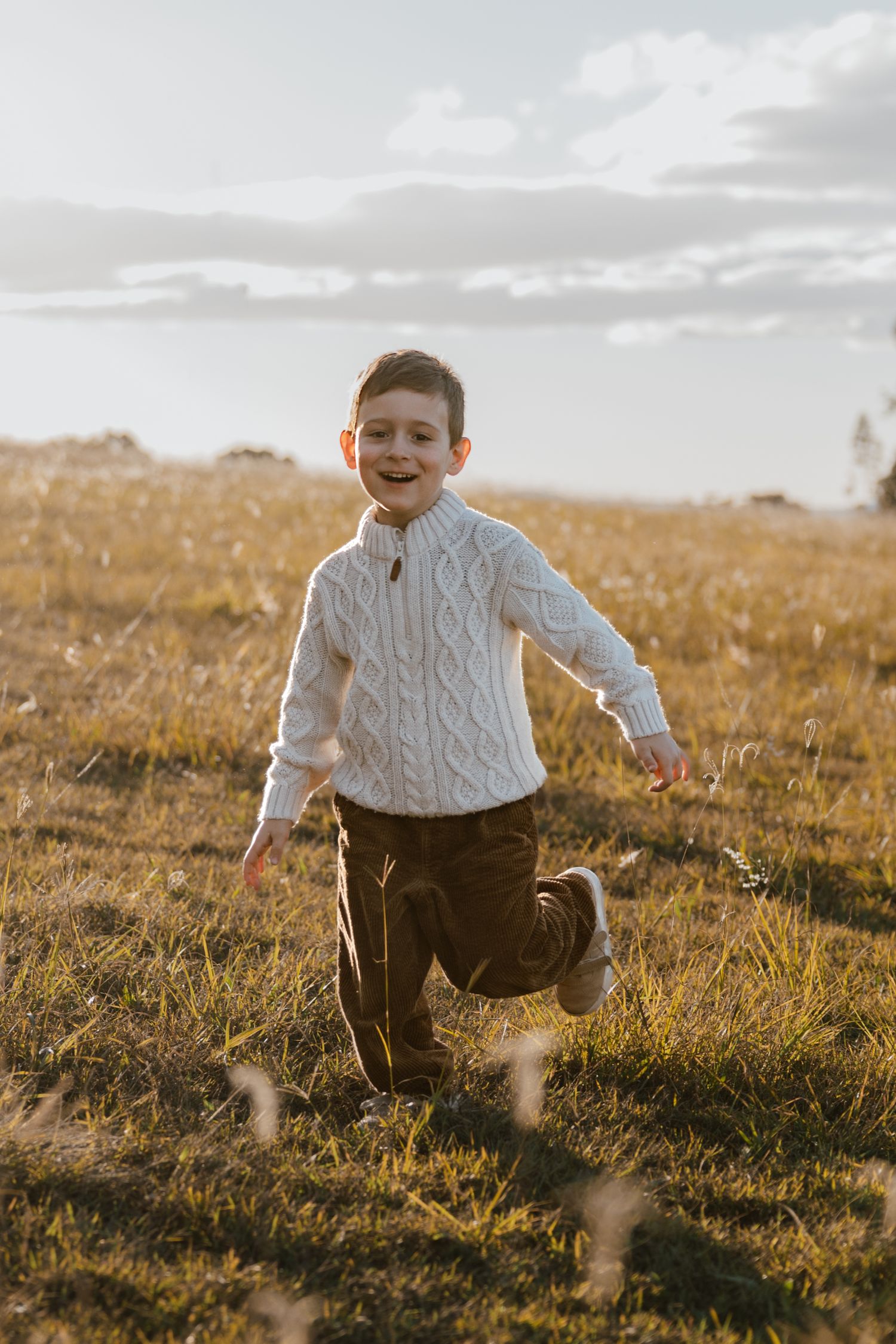 A smiling young boy in a cream cable knit sweater and brown corduroy pants runs through a grassy field under a soft evening sky.