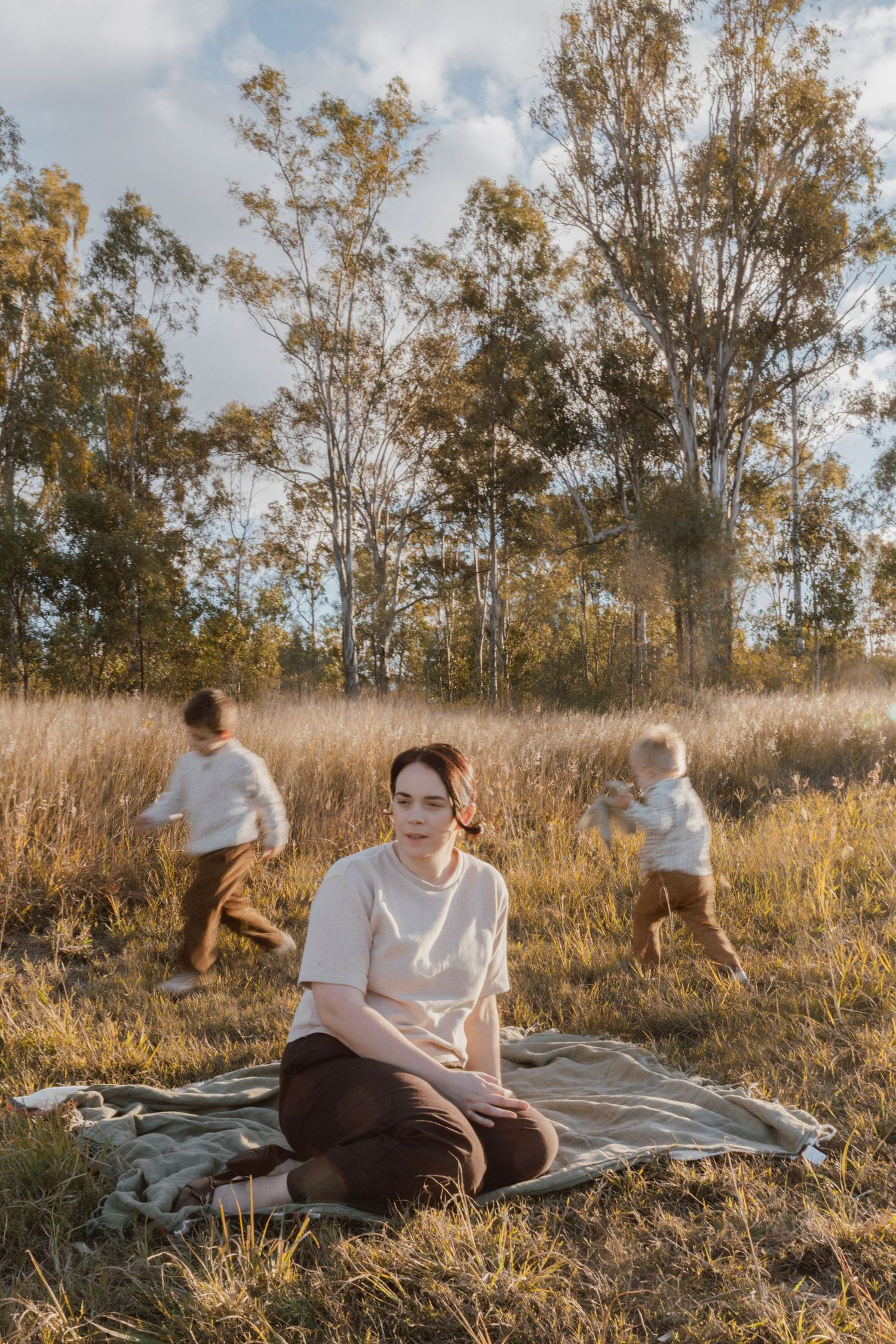 A mother sits on a blanket in a golden grass field while her two young sons play behind her among tall gum trees at sunset.