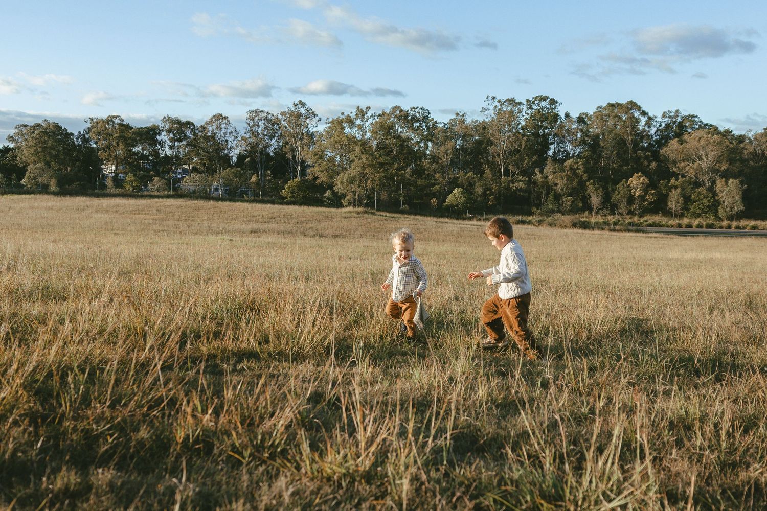 Two small boys run through tall golden grass in an open field with a line of trees under a soft blue sky.