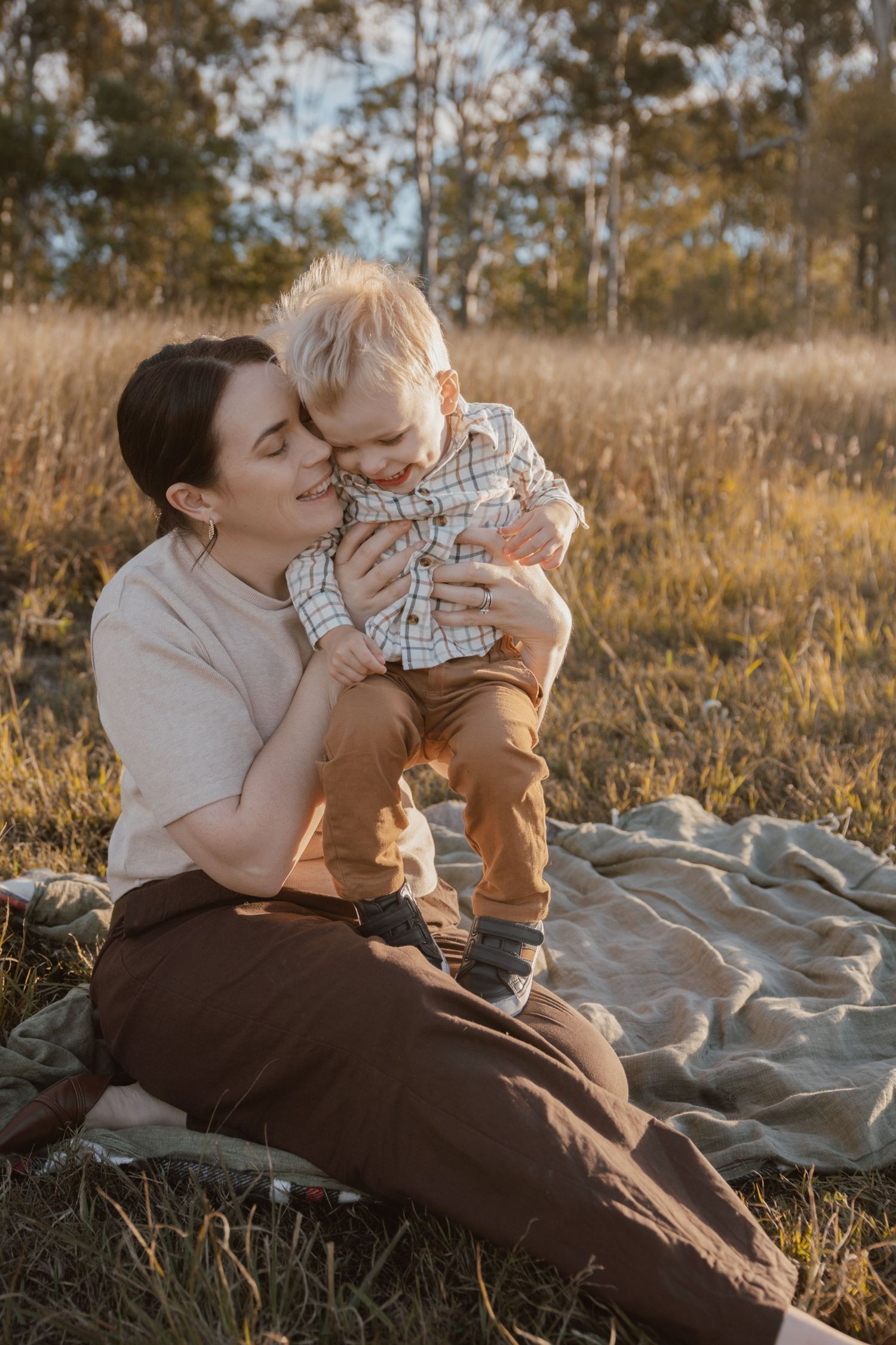 A mother in a cream top laughs and hugs her blond toddler son on a blanket in a golden grassy field surrounded by trees.
