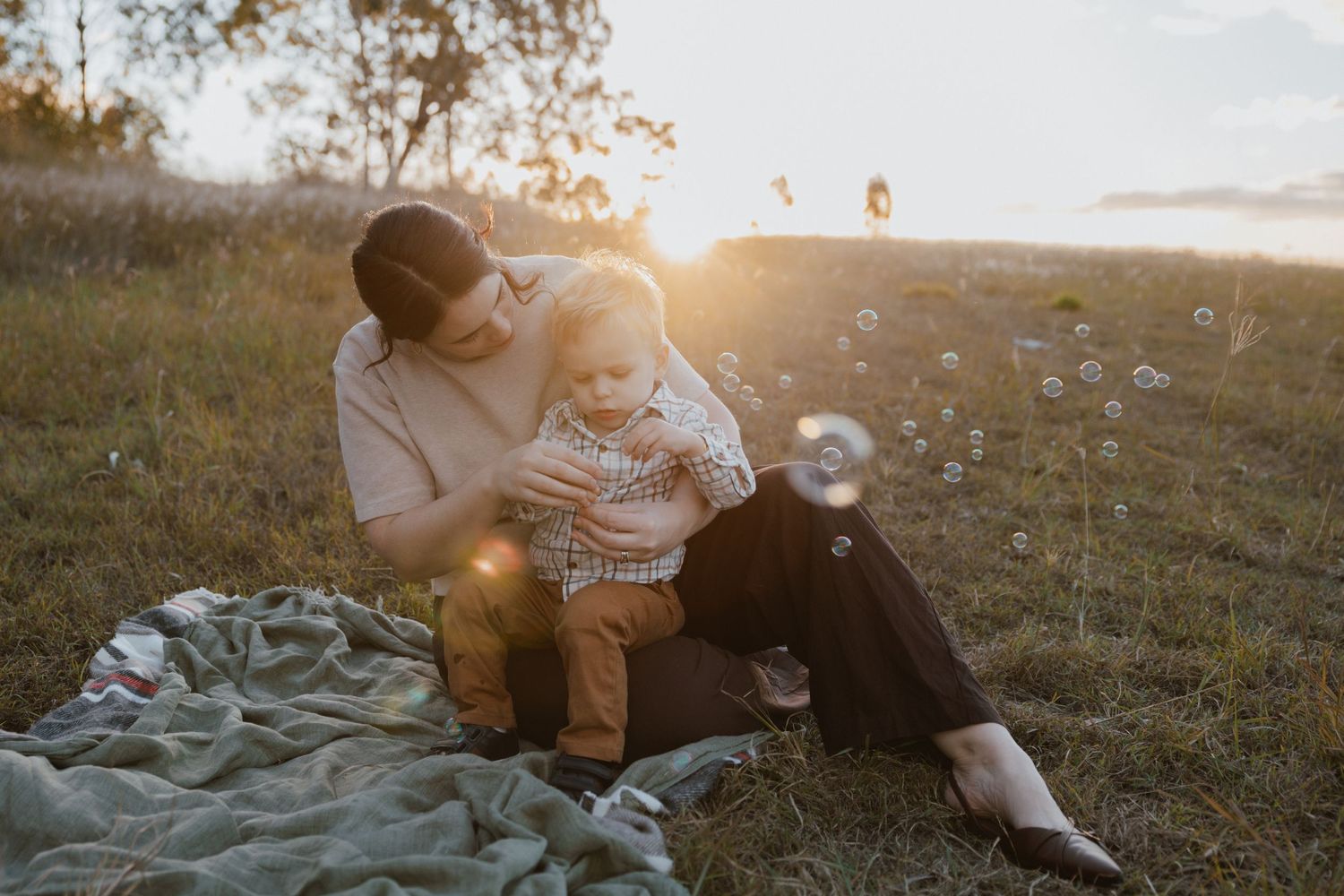 A mother cuddles her toddler son on a blanket in a sunlit field as soap bubbles float through the warm golden hour light.