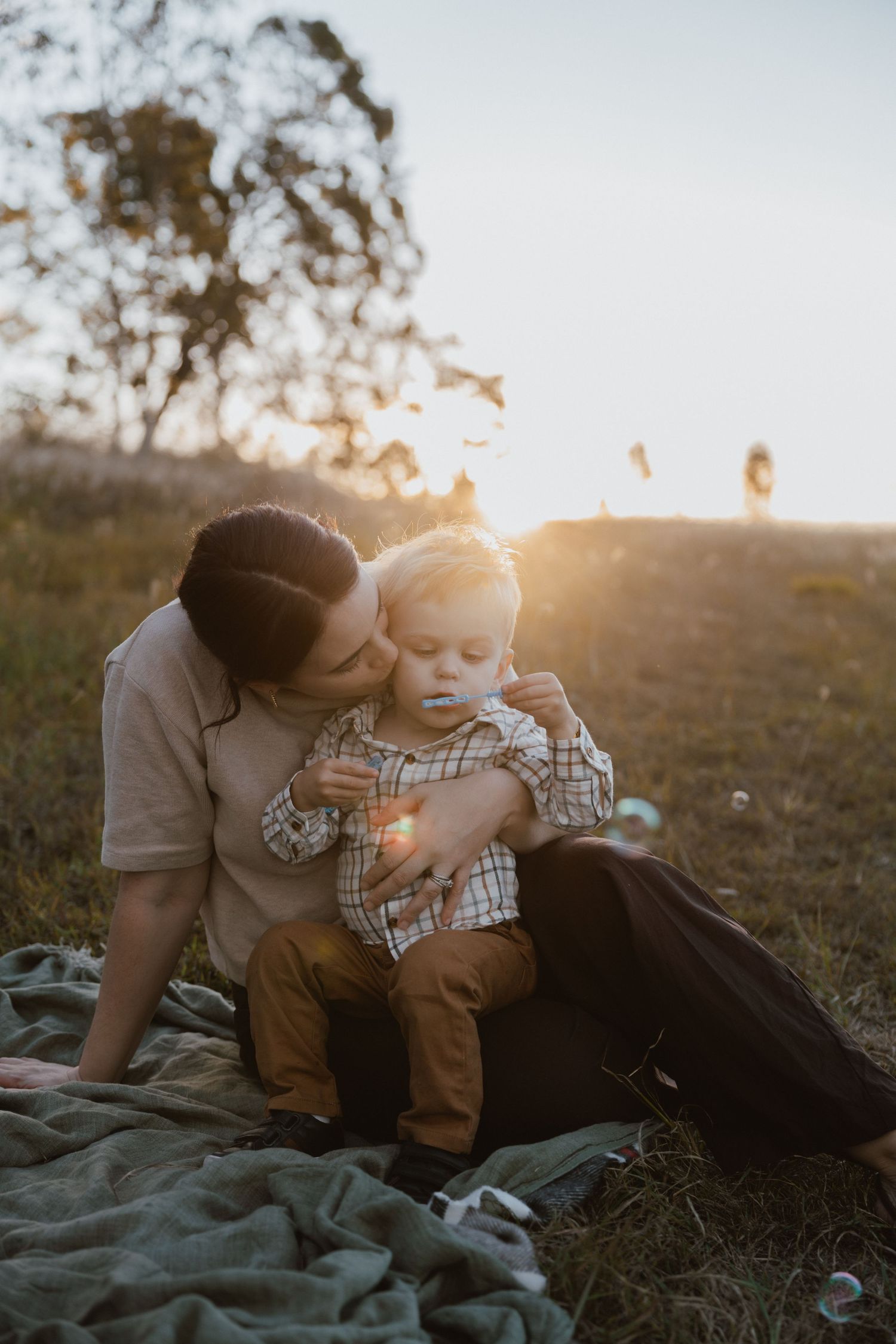 A mother kisses her toddler son on the cheek while sitting on a blanket in a sunlit meadow as soap bubbles float nearby.