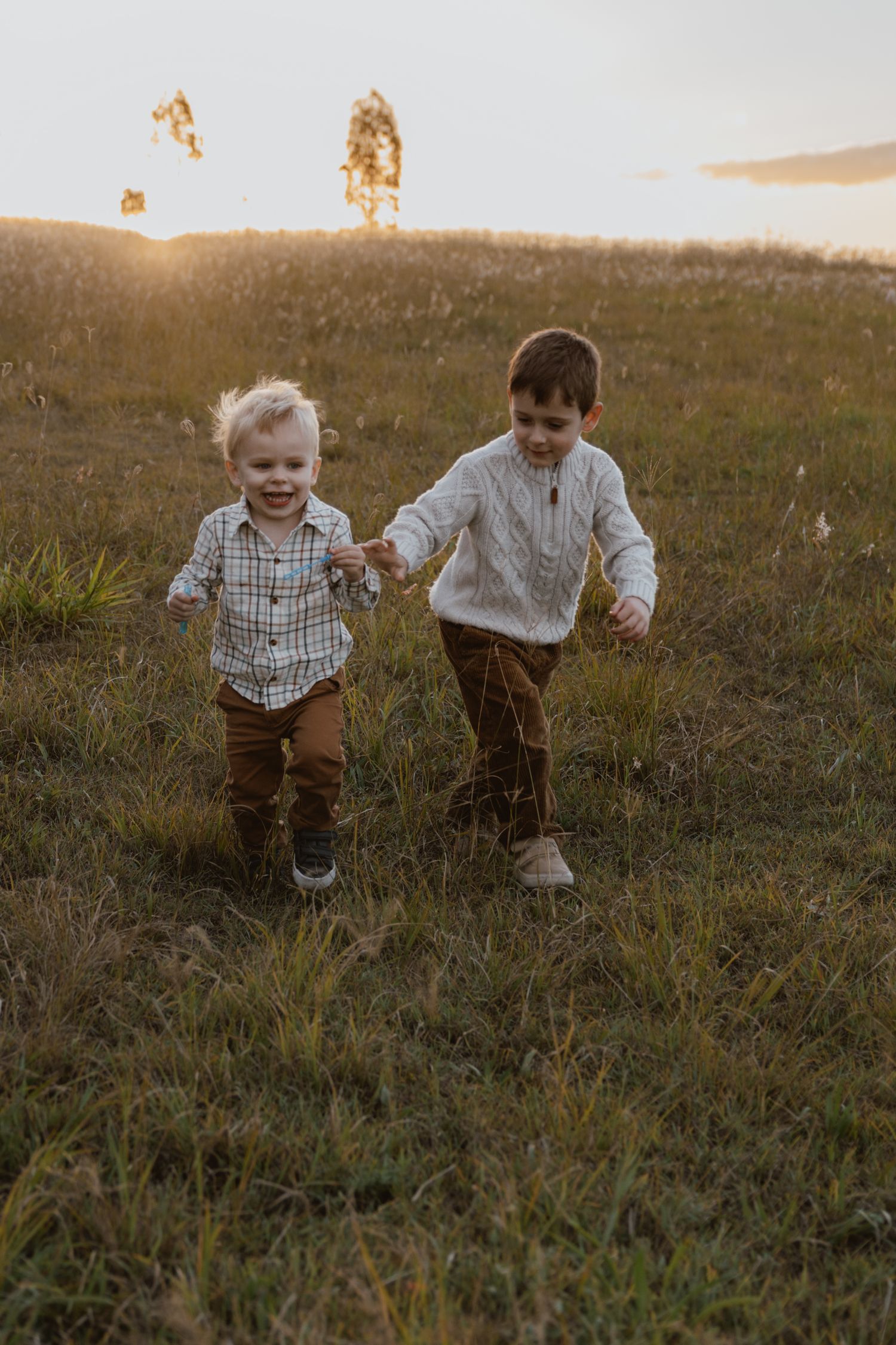 Two young brothers in coordinating outfits hold hands and run joyfully through a grassy field at golden hour sunset.