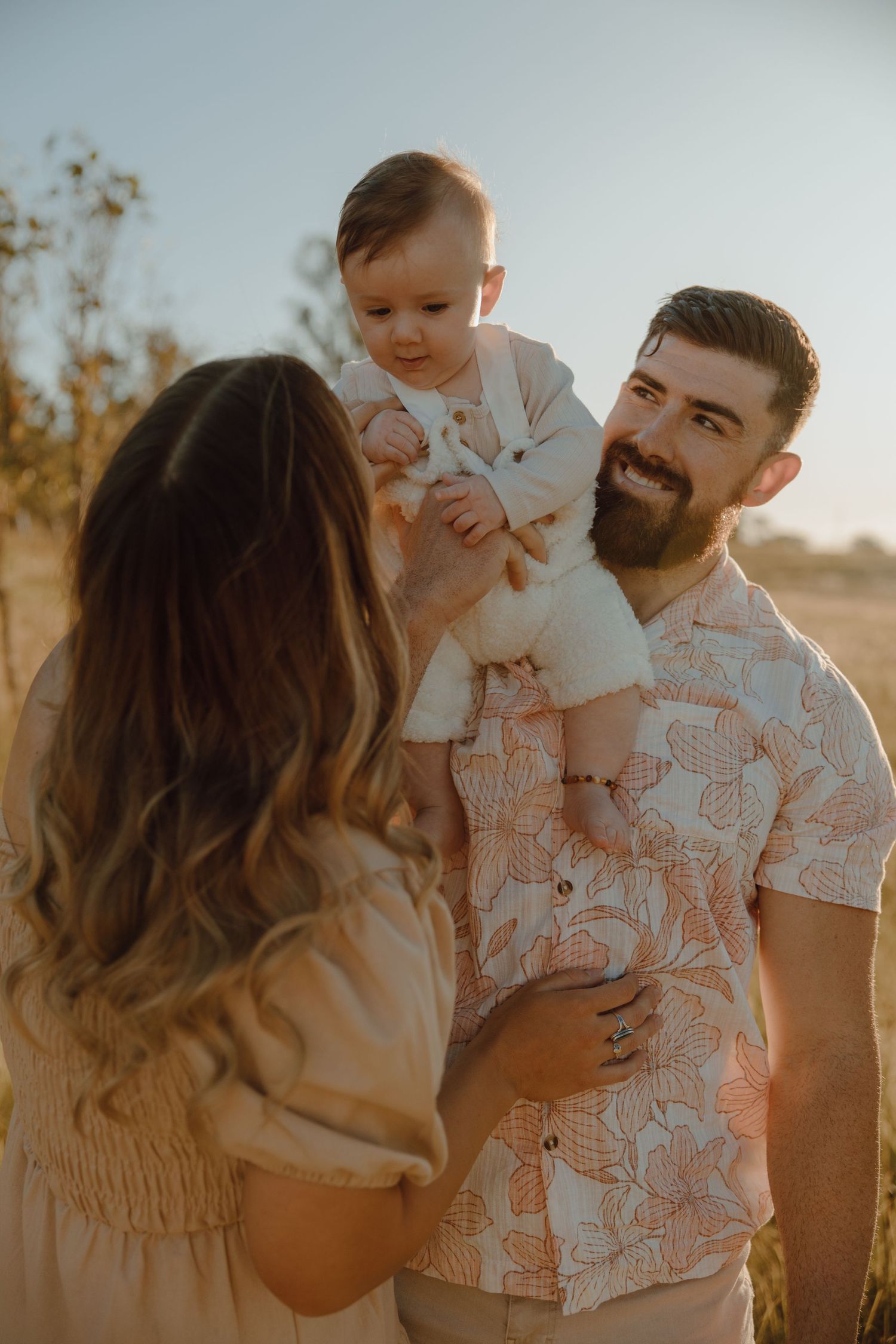 A bearded father in a floral shirt lifts his smiling baby into the air while the mother looks on in golden sunset light.