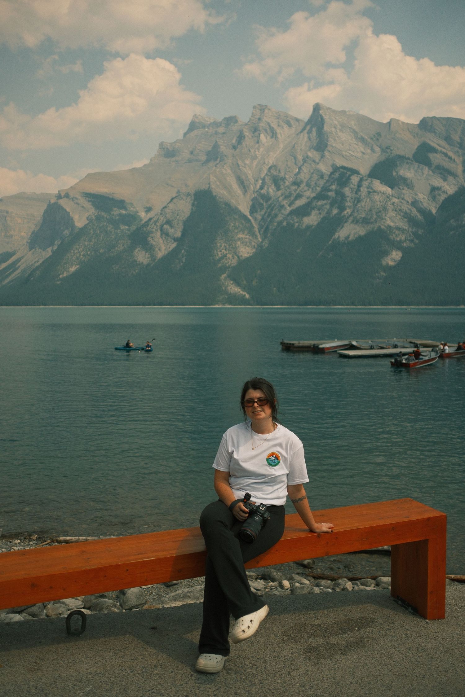 Woman in sunglasses and white tee sits on an orange bench beside a turquoise mountain lake with kayakers and rocky peaks.