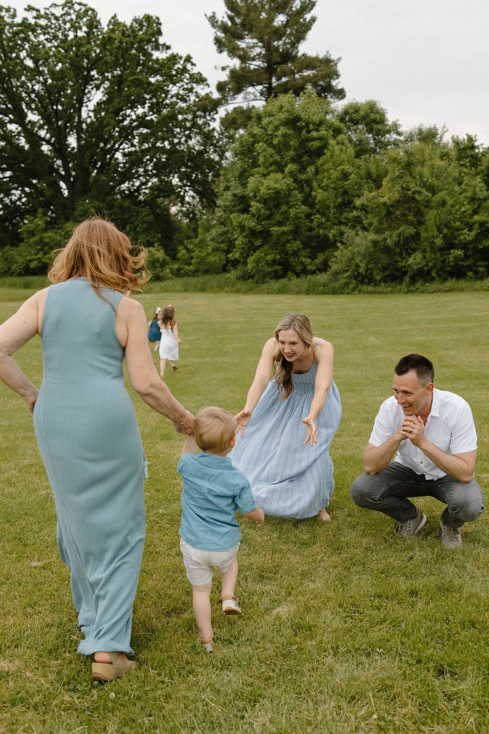 A family shares a playful moment together on a grassy lawn surrounded by trees on a summer day.