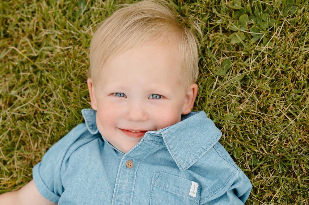 A toddler wearing a light blue denim shirt lies on green grass with a cheerful smile.